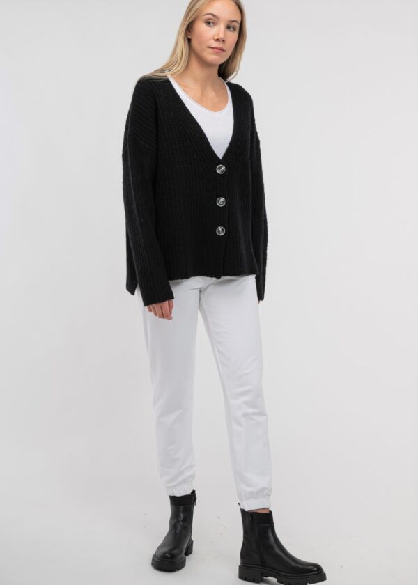 KAYA 100% Wool V- Ausschnitt Strickjacke in schwarz, in einer grobgestrickten Patent Rippe mit knöpfen und überschnittener Schulter an blonden jungen frau mit weißer Hose und Top, von vorne