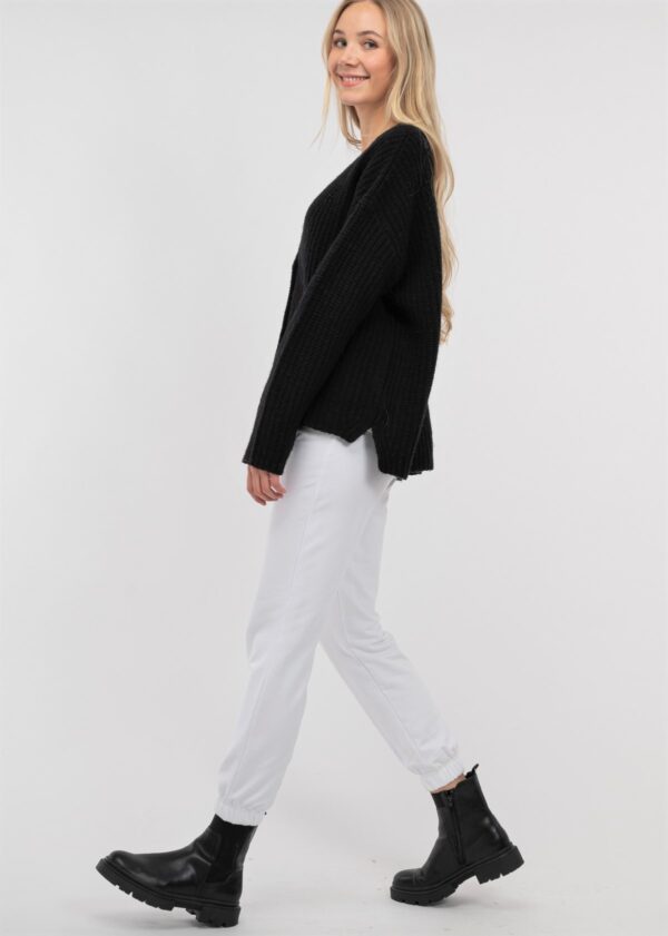 KAYA 100% Wool V- Ausschnitt Strickjacke in schwarz, in einer grobgestrickten Patent Rippe mit knöpfen und überschnittener Schulter an blonden jungen frau mit weißer Hose und Top, von der Seite
